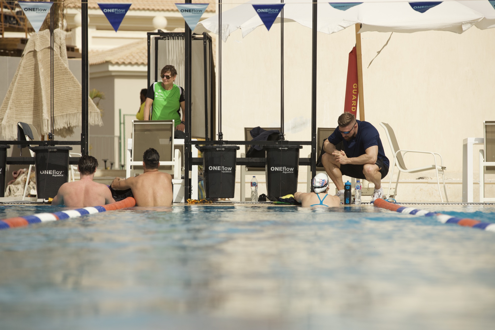 Coach am Beckenrand im Gespräch mit zwei erwachsenen Schwimmern in der Anlage von ONEflow Sports in Soma Bay